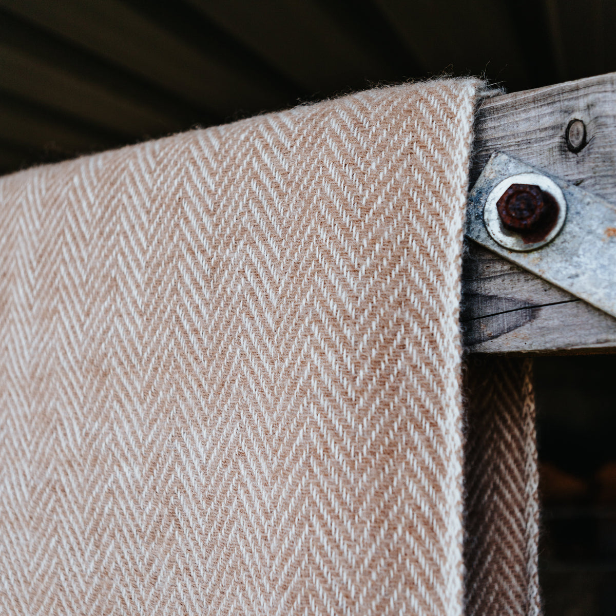 A close-up of a fawn and cream herringbone woven alpaca wool textile, draped over a rustic wooden post with a metal grommet.