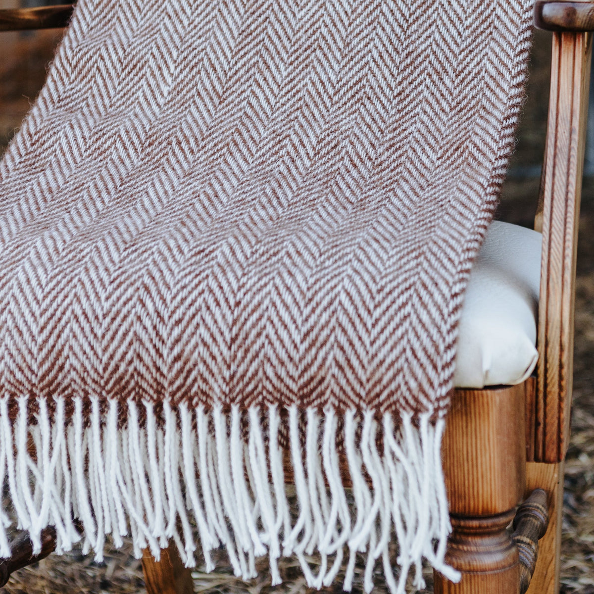 A woven throw blanket with a brown and cream herringbone pattern and white fringe, draped over a wooden chair in a barn setting.