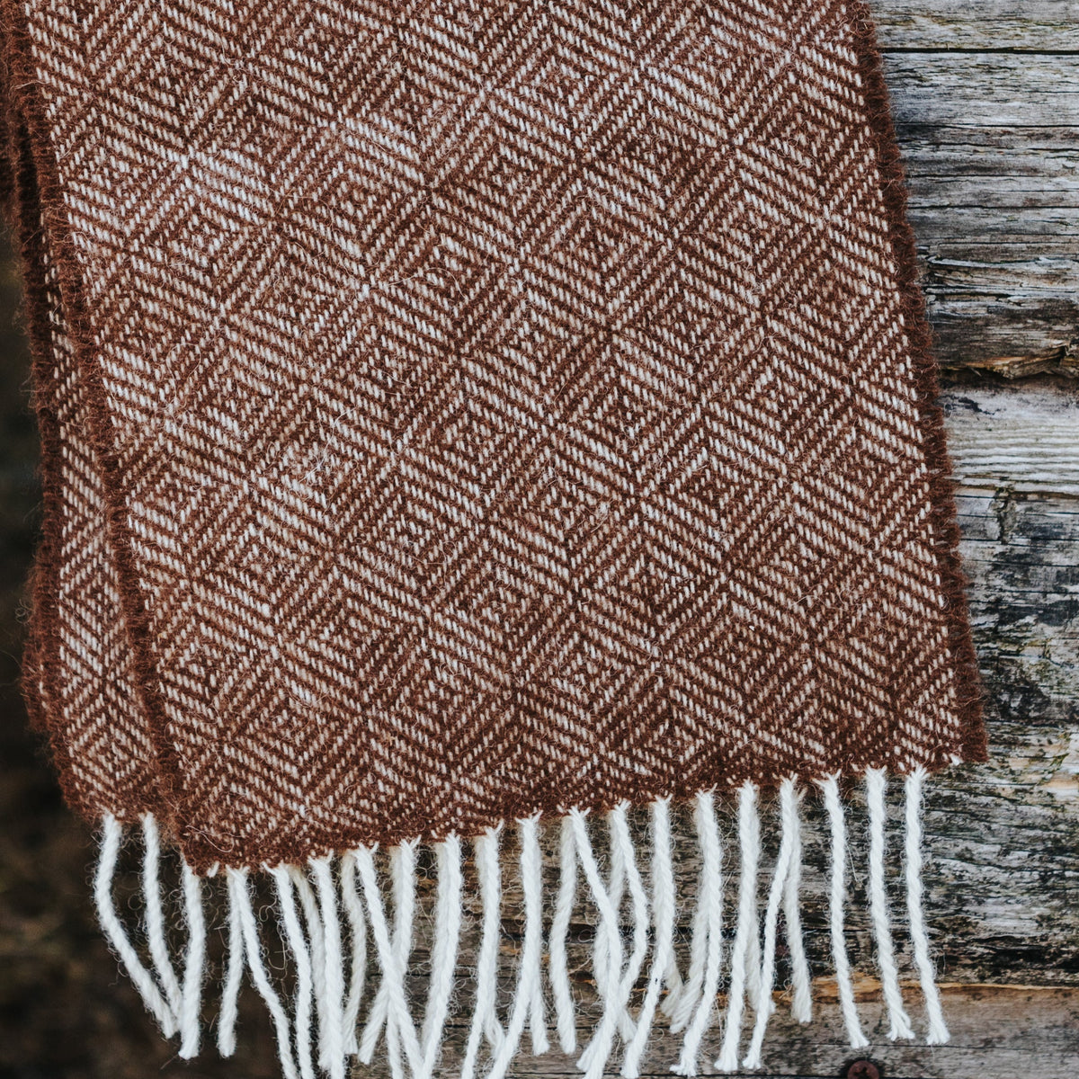 A brown and cream herringbone woven alpaca textile with white fringe, draped over a rustic wooden post in a barn setting.