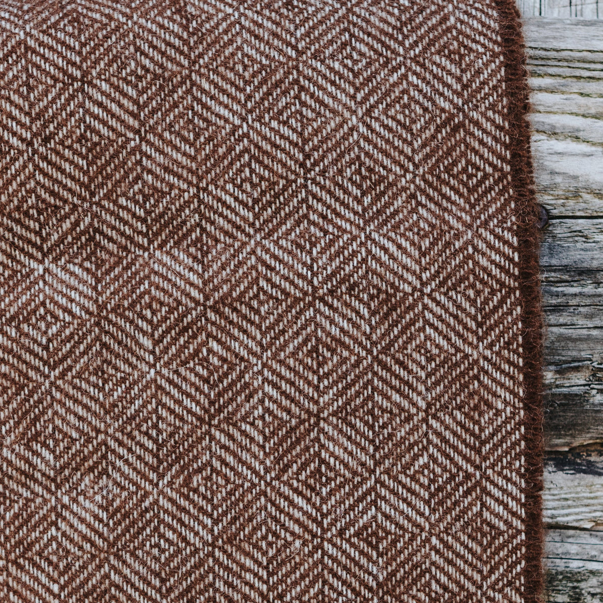 A brown and cream diamond woven alpaca textile, hanging on a rustic weathered wooden post in a barn setting.