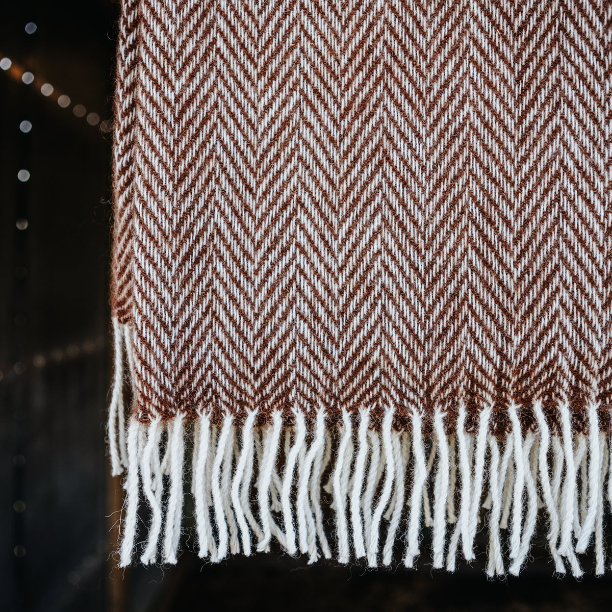 A close-up of a woven throw blanket with a brown and cream herringbone pattern and white fringe tassels, hanging against a dark background with soft string lights.