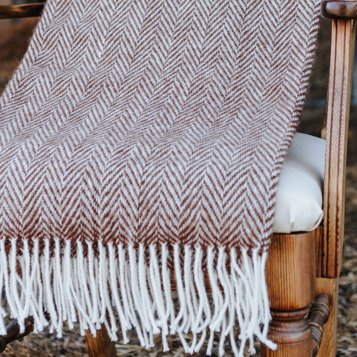 A woven throw blanket with a brown and cream herringbone pattern and white fringe, draped over a wooden chair in a barn setting.