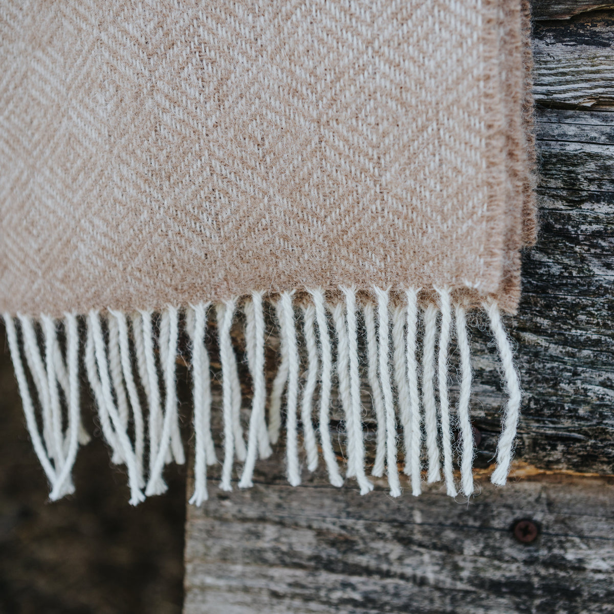 A close-up of a woven scarf in a fawn and cream herringbone pattern with white fringe, draped over a rustic dark wood surface.