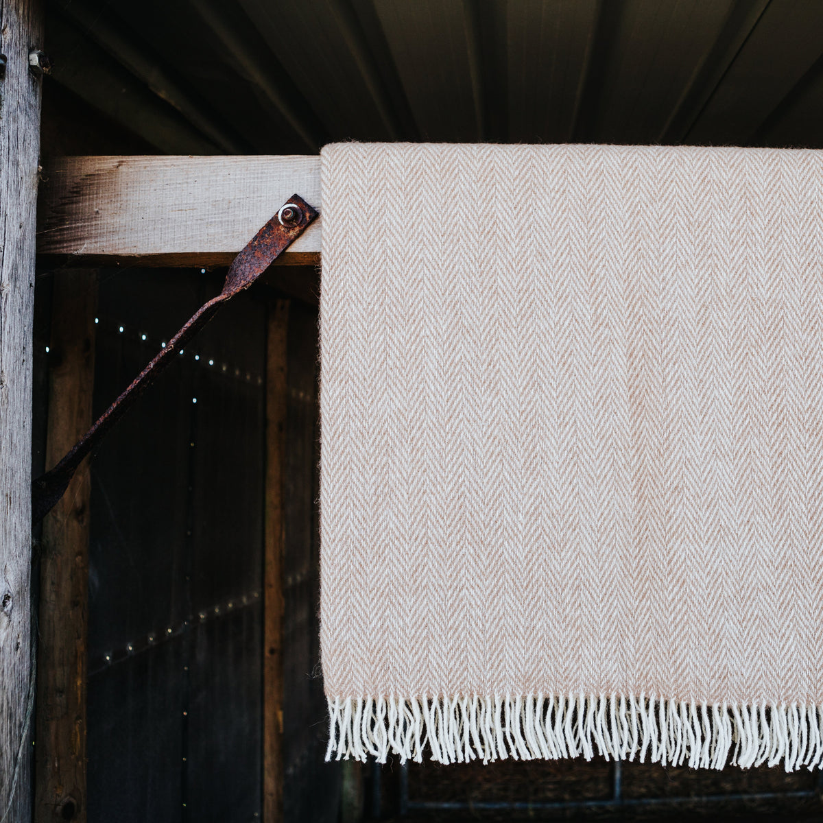 A fawn cream herringbone alpaca woven blanket with white fringe hanging from a rustic wooden barn door.