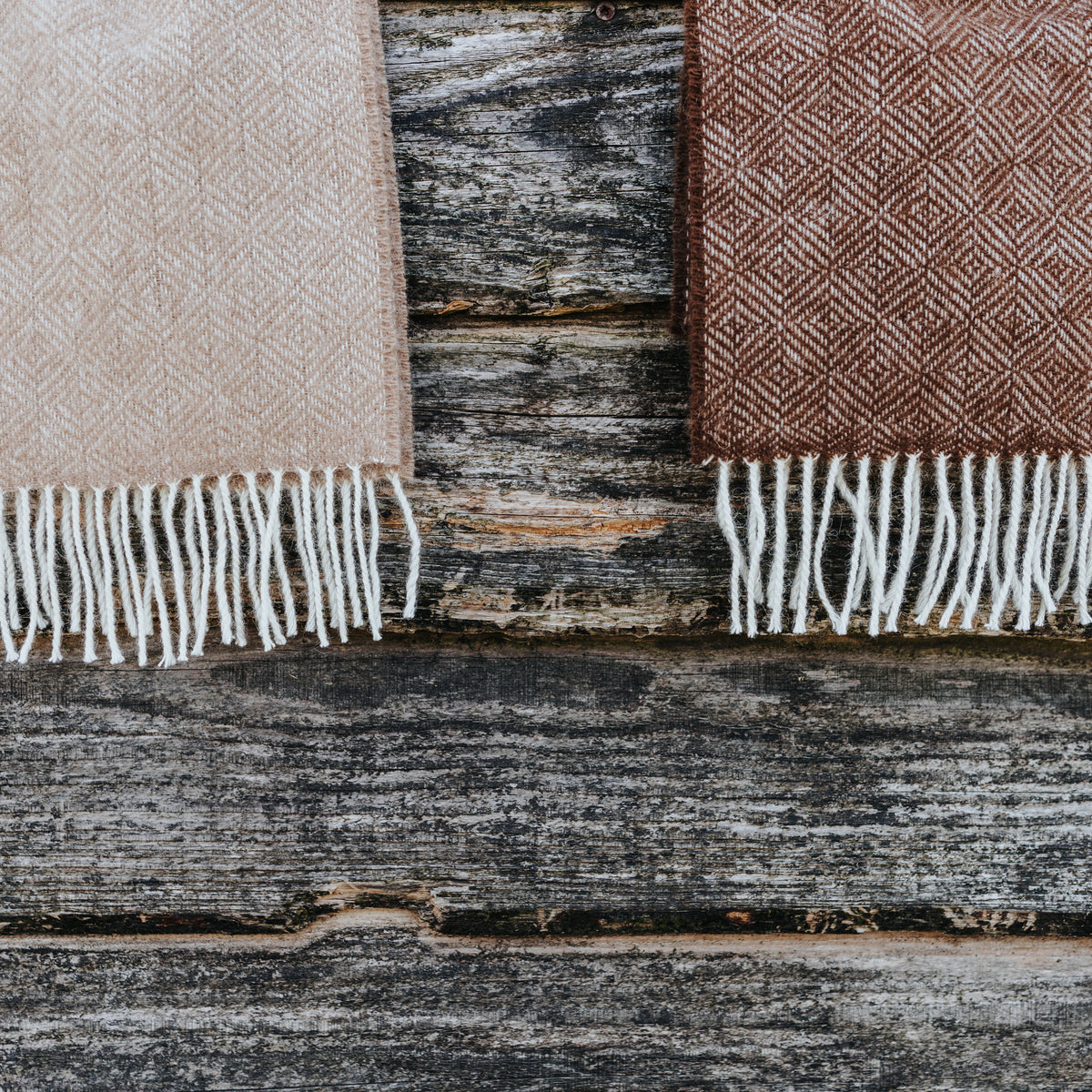 Two small-batch luxury Rhynaston Alpaca wool scarves in natural fawn herringbone and dark chocolate diamond weave, folded side-by-side on a rustic weathered barn wood background in Pembrokeshire, Wales.